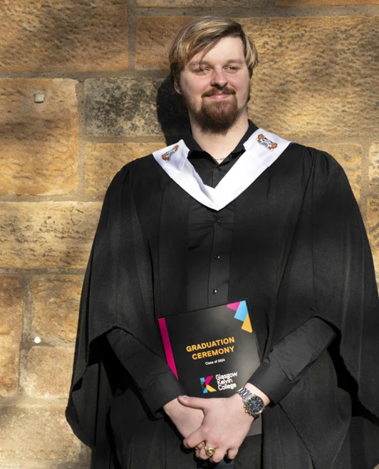Male graduate in gown posing in front of a stone wall, holding a graduation certificate with a confident smile. Male graduate in gown posing in front of a stone wall, holding a graduation certificate with a confident smile.